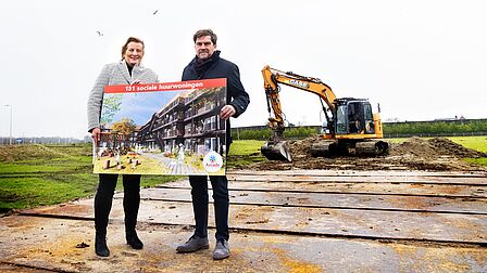Wethouder Carlieke van Staalduinen en directeur-bestuurder Wilfried Stribos staan voor een graafmachine en houden een poster vast over sociale huurwoningen.